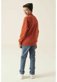 Sudadera naranja, vaqueros azules, zapatillas blancas con el logo de "Nike", gorro negro. El modelo está girado, mostrando un atuendo casual.