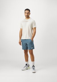 Light beige athletic t-shirt with a dark accent stripe, paired with blue shorts featuring side pockets and a white logo, completed with white sneakers.