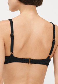 Čierny bikiny top s rebrovaným vzorom, ktorý má nastaviteľné popruhy, zapínanie na háčik a textúrovaný povrch, zdôrazňujúci jednoduchosť a funkčnosť.