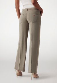 Pantaloni largi de culoare khaki deschis, realizați dintr-un material moale, având două buzunare pe spate și un mic etichetă cu logo. Asortati cu sandale cu toc înalt de culoare bej.