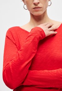 Egy nő vörös, hosszú ujjú felsőt visel, egyik kezét a kulcscsontjára támasztja, és fülbevalói jól látszanak egy egyszínű háttér előtt.