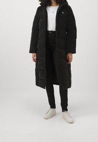 Person som bär en lång svart vadderad kappa, vit skjorta med logga, svarta byxor och vita platåsneakers, stående mot en vanlig bakgrund.