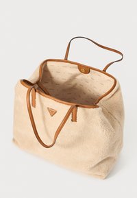 Beige faux fur tote bag med brune læderhåndtag og et trekantet logo. Indvendigt har den en tan-farvet foring med et gentaget "GUESS" mønster.
