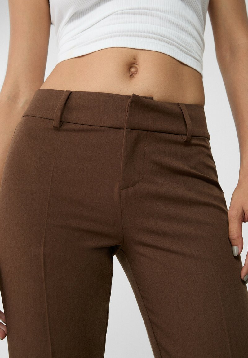 Pantaloni marroni su misura con una texture liscia, dotati di vita piatta, passanti per la cintura e tasche laterali. La vestibilità è affusolata e strutturata.