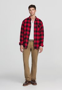 Giovane uomo in piedi, che indossa una camicia a quadri rossi e neri sopra una t-shirt bianca, pantaloni beige, scarpe marroni e un'espressione neutra su uno sfondo grigio.