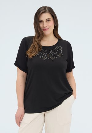 Kvinde med langt brunt hår iført en sort kortærmet top med blomsterblonde-detalje og lyse beige bukser, stående med den ene hånd i lommen.