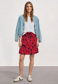 Femme debout portant une veste bleu clair, une chemise blanche, une jupe rouge fleurie et des baskets beiges avec des bandes bordeaux sur un fond uni.