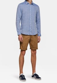 Světle modrá lněná košile na knoflíky, světlé krátké kalhoty a námořnické tenisky. Košile má jemnou texturu; kraťasy mají klasický střih.