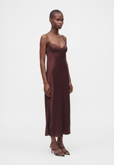 Femme mince portant une robe en satin marron foncé sans manches avec de fines bretelles et des sandales noires à talons, debout devant un fond clair uni.