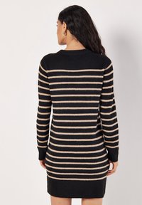 Femme aux longs cheveux foncés portant une robe-pull à manches longues à rayures horizontales noires et beiges, vue de dos sur un fond blanc.