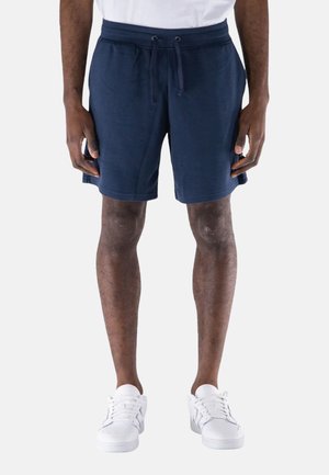 Pantaloni corti blu navy realizzati in un tessuto morbido, con vita regolabile con coulisse e tasche laterali. Indossati con sneakers bianche.