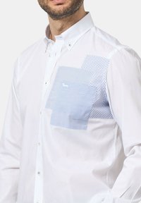 Camicia bianca con bottoni, caratterizzata da accenti blu a motivo sul petto, con diverse forme e texture per un contrasto visivo.
