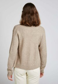 Pull en tricot beige avec des côtes verticales, encolure ronde et épaules légèrement tombantes. Texture douce et coupe décontractée, associé à un pantalon léger.