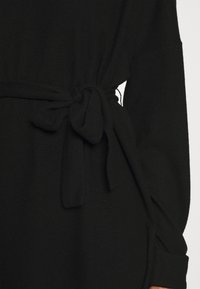 Svart stickad plagg med knytbart midjebälte, med långa ärmar och en texturerad yta, som betonar en avslappnad passform och minimalistisk design.