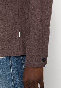 Hnědá košile z vlněné směsi s texturovaným povrchem, s knoflíkovým manžetem a nenápadnou logo štítkem na boku. Kombinována s modrými džínami.