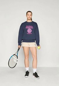 Jauna moteris, vilkinti tamsiai mėlyną džemperį ir béžinius šortus, viena ranka laiko teniso raketę, o kita – teniso kamuoliuką, stovinti ant vienodo fono.