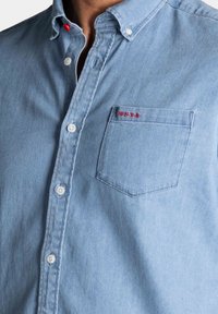Lichtblauwe spijkerblouse met button-down kraag, witte knopen, borstzak en rode geborduurde logodetail op de zak.