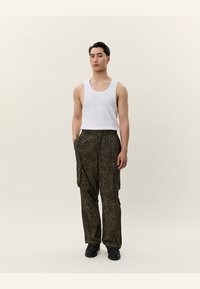 Ținută bărbații compusă dintr-un maiou alb și pantaloni cargo verzi în camuflaj închis, cu multiple buzunare, confecționați dintr-un material ușor. Pantofii negri completează aspectul.
