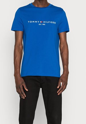 Mand iført en blå Tommy Hilfiger t-shirt med logo og teksten "EST. 1985", kombineret med sorte bukser mod en ensfarvet baggrund.