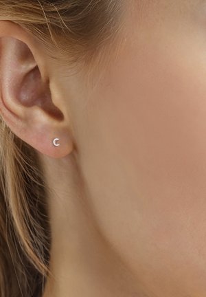 Nahaufnahme eines Frauenohrs mit kleinem, silbernem, halbmondförmigem Ohrstecker und hellbraunem Haar, das hinter dem Ohr zurückgesteckt ist.