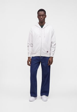 Ein Mann trägt einen weißen Kapuzenpullover mit Reißverschluss, blaue Jeans mit weitem Bein und weiße Turnschuhe und steht vor einem schlichten weißen Hintergrund.