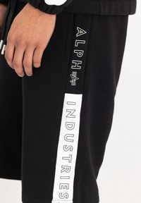 Shorts noirs en mélange de coton, avec une accentuation blanche portant le texte "ALPHA INDUSTRIES" sur le côté. Texture lisse, coupe décontractée.