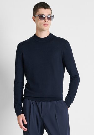 Junger Mann mit dunkler Sonnenbrille, einem enganliegenden schwarzen Langarmpullover und dunklen plissierten Hosen steht mit einer Hand in der Tasche.