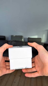 Witte rechthoekige doos met een groen Lacoste-logo bovenop en aan de voorkant, vastgehouden tussen twee handen, glad oppervlak, minimalistisch ontwerp.