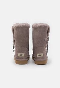 Bottines en daim gris avec une bordure rose duveteuse, semelle plate et étiquette UGG à l'arrière. Présentent un accent décoratif sur le côté.