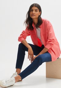 Korallfarbenes Button-Down-Hemd mit zwei vorderen Taschen, kombiniert mit dunklen Jeans und weißen Sneakern. Glattes Material mit hochgekrempelten Ärmeln.