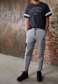 Šedé tepláky s bočními kapsami, černými akcenty a stahovací šňůrkou, v kombinaci s námořnickým tričkem Nike s bílými pruhovanými rukávy. Černé tenisky.