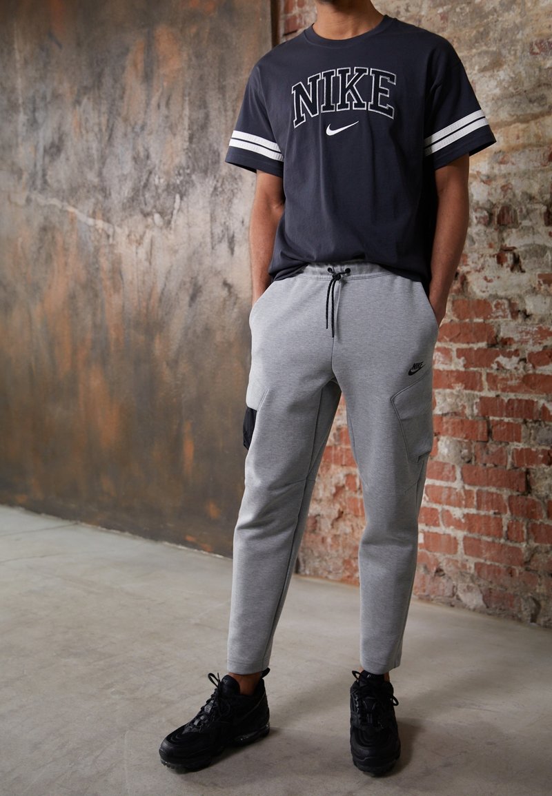 Šedé tepláky s bočními kapsami, černými akcenty a stahovací šňůrkou, v kombinaci s námořnickým tričkem Nike s bílými pruhovanými rukávy. Černé tenisky.