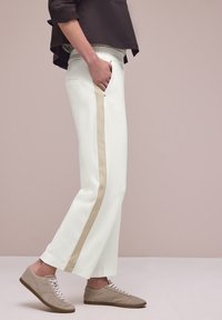 Vue de profil d'une personne portant un pantalon blanc avec une bande latérale beige, un haut marron et des chaussures décontractées beiges à lacets, debout avec une main dans la poche.