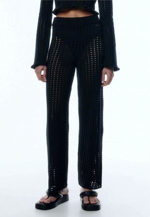 Pantalon en maille crochet noir porté avec un top court assorti et des sandales plateformes noires sur un mannequin se tenant devant un fond blanc.