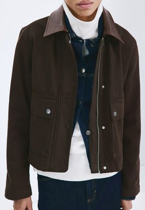 Persona con ropa en capas, lleva una chaqueta marrón oscura sobre una chaqueta vaquera azul oscuro y un suéter de cuello alto blanco con textura acanalada.