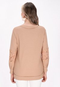 Beige kötött pulóver, hosszú ujjakra virágos csipkakibővítéssel. Bordás textúra, kerek nyakkivágás és laza fazon.