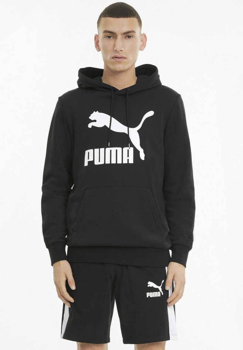 Svart bomullshettegenser med kengurulomme, med et stort hvitt Puma-logo. Kombinert med svarte shorts som også har Puma-logo i hvitt.
