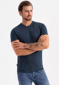 Námořnická modrá tričko s kulatým výstřihem z hladké tkaniny, s malým logem na prsou. V kombinaci s modrými džínami.
