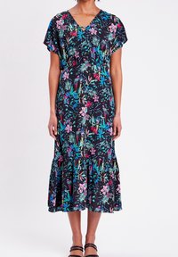 Femme portant une robe midi noire avec des imprimés floraux et d'oiseaux vifs, des manches courtes, un décolleté en V, un ourlet volanté et des sandales noires à bride chevillère.
