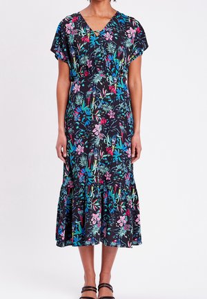 Femme portant une robe midi noire avec des imprimés floraux et d'oiseaux vifs, des manches courtes, un décolleté en V, un ourlet volanté et des sandales noires à bride chevillère.