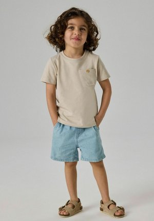 Copil cu păr creț purtând un tricou bej, pantaloni scurți bleu și sandale maro, stând cu mâinile în buzunare pe un fundal simplu.