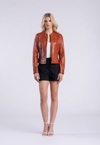 Bruine leren jacket met een mandarin kraag, twee voorzijde ritszakken en een getailleerd ontwerp, gecombineerd met zwarte shorts en witte gestreepte sandalen.