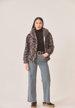 Mujer de pie con una chaqueta de pelo sintético con estampado de leopardo, cuello alto negro, vaqueros grises de pierna ancha, pendientes de aro dorados y zapatillas negras.