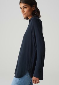 Blusa a maniche lunghe blu navale con una vestibilità rilassata, dotata di un colletto alto, polsini con bottoni e un tessuto morbido e liscio. L'orlo è leggermente arrotondato.