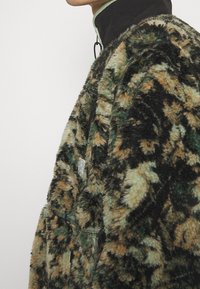 Fleecejacke in Grün, Braun und schwarzem Blumenmuster; verfügt über eine Reißverschlusstasche, hohen Kragen und weichen Stoff.