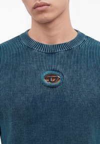 Gros plan d'une personne portant un pull côtelé bleu avec un petit logo métallique ovale centré sur la poitrine.