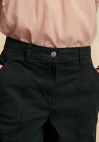 Des pantalons en mélange de coton noir qui présentent une taille ajustée avec un bouton en métal argenté et deux poches avant. Le tissu a une texture lisse avec des coutures visibles.