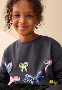 Temno siva majica z okroglim izrezom, okrašena s pisanimi metulji in pentljami iz cekinov v roza, modri in rumeni barvi. Mehka tkanina.