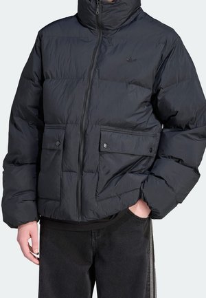 Zwarte pufferjas met ritssluiting aan de voorkant en twee klepzakken, gedragen met één hand in de zak en een subtiel logo op de borst.