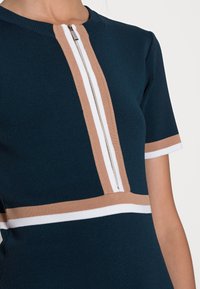 Robe en maille bleu foncé à manches courtes, dotée d'une fermeture éclair centrale et de rayures horizontales beige et blanches sur le torse.